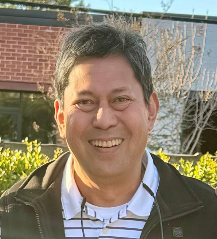 A man wearing a black jacket and a striped shirt is smiling for the camera.