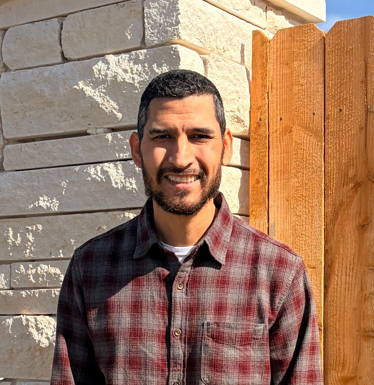 Man with a beard smiling, wearing a red plaid shirt, standing in front of a stone wall and wooden fence.