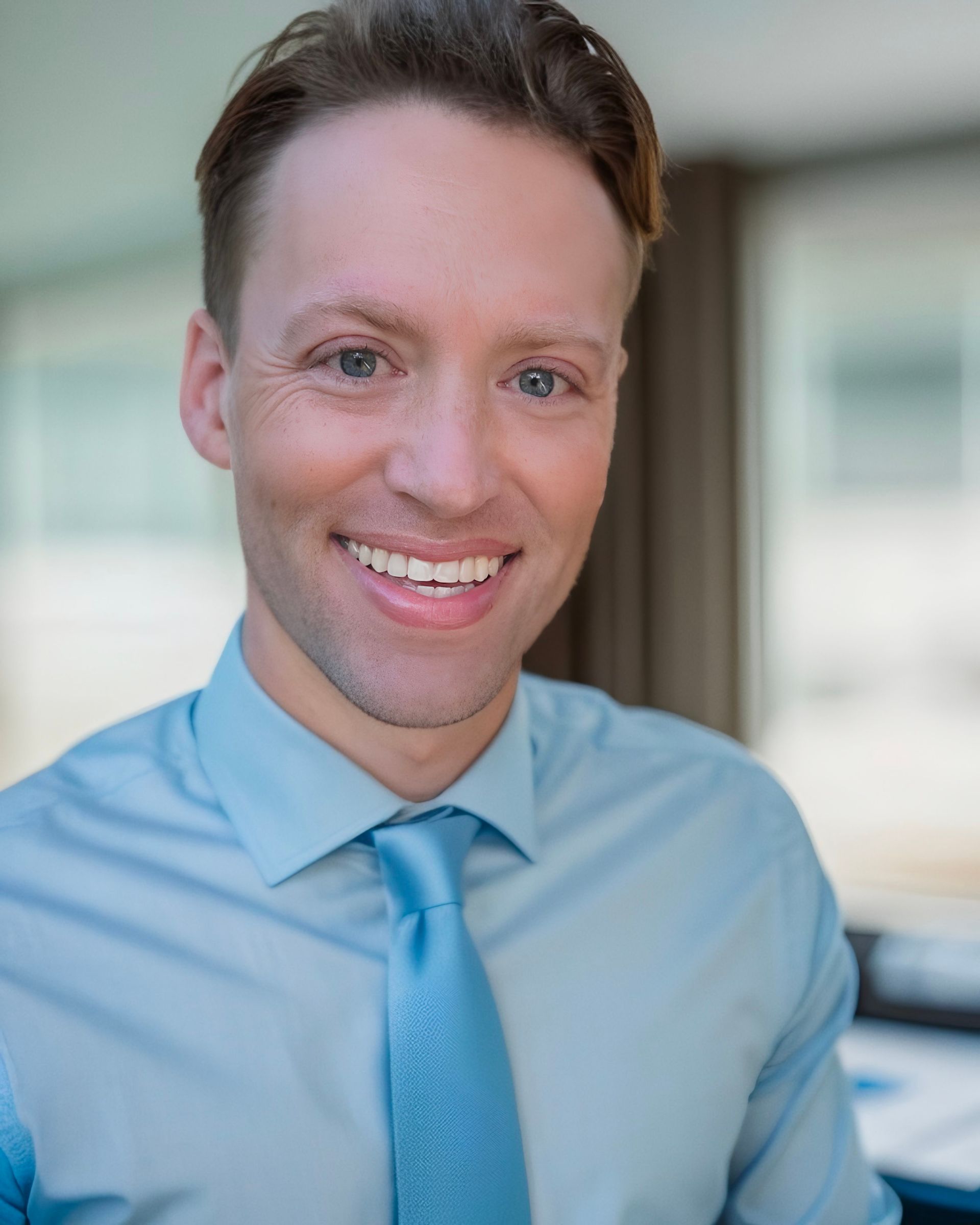 Man with light hair and blue tie smiles at the camera, wearing a light blue shirt.