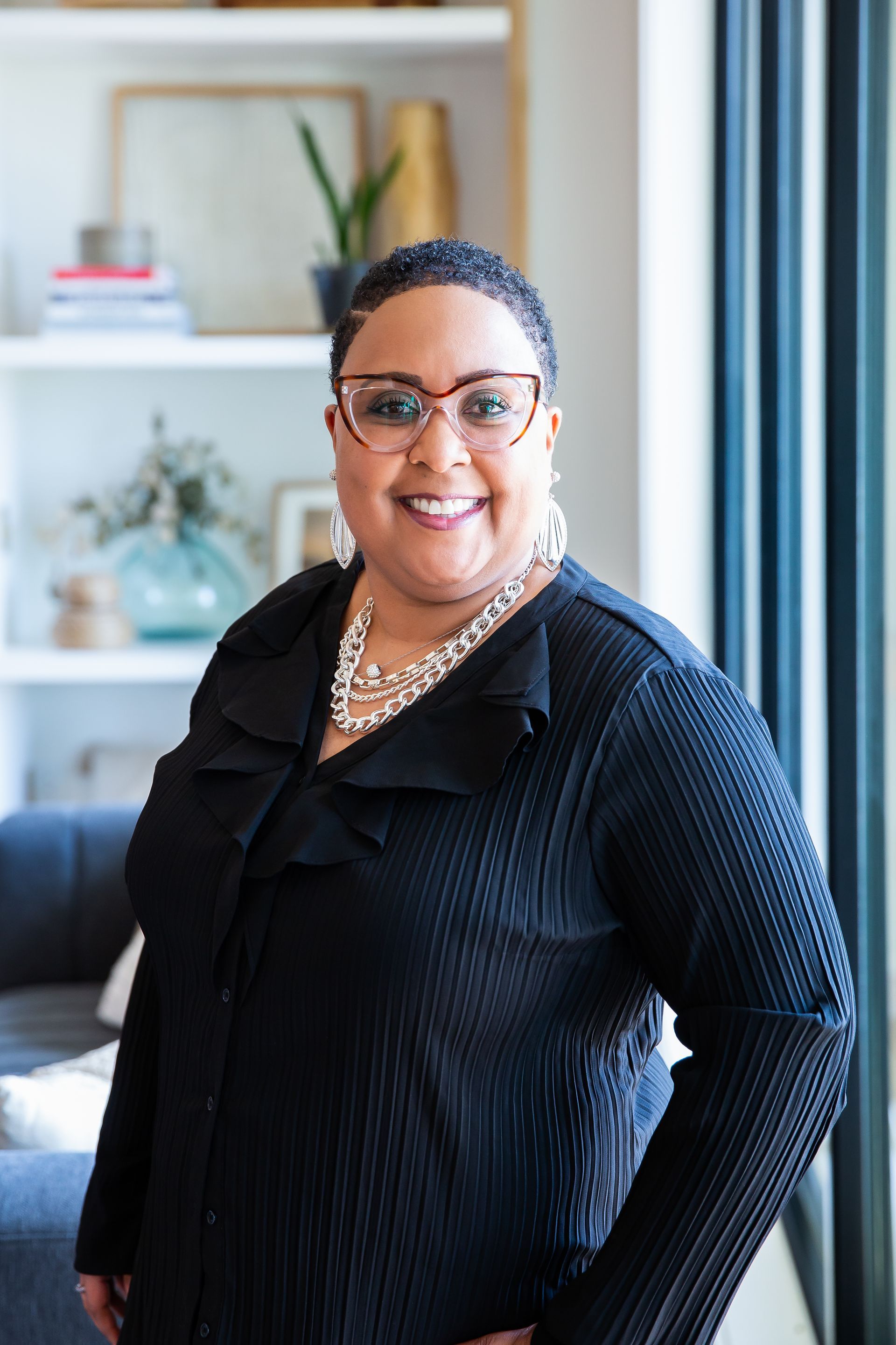 A woman wearing glasses and a necklace is standing in front of a window in a living room.