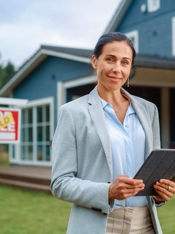 A woman is standing in front of a blue house holding a tablet.