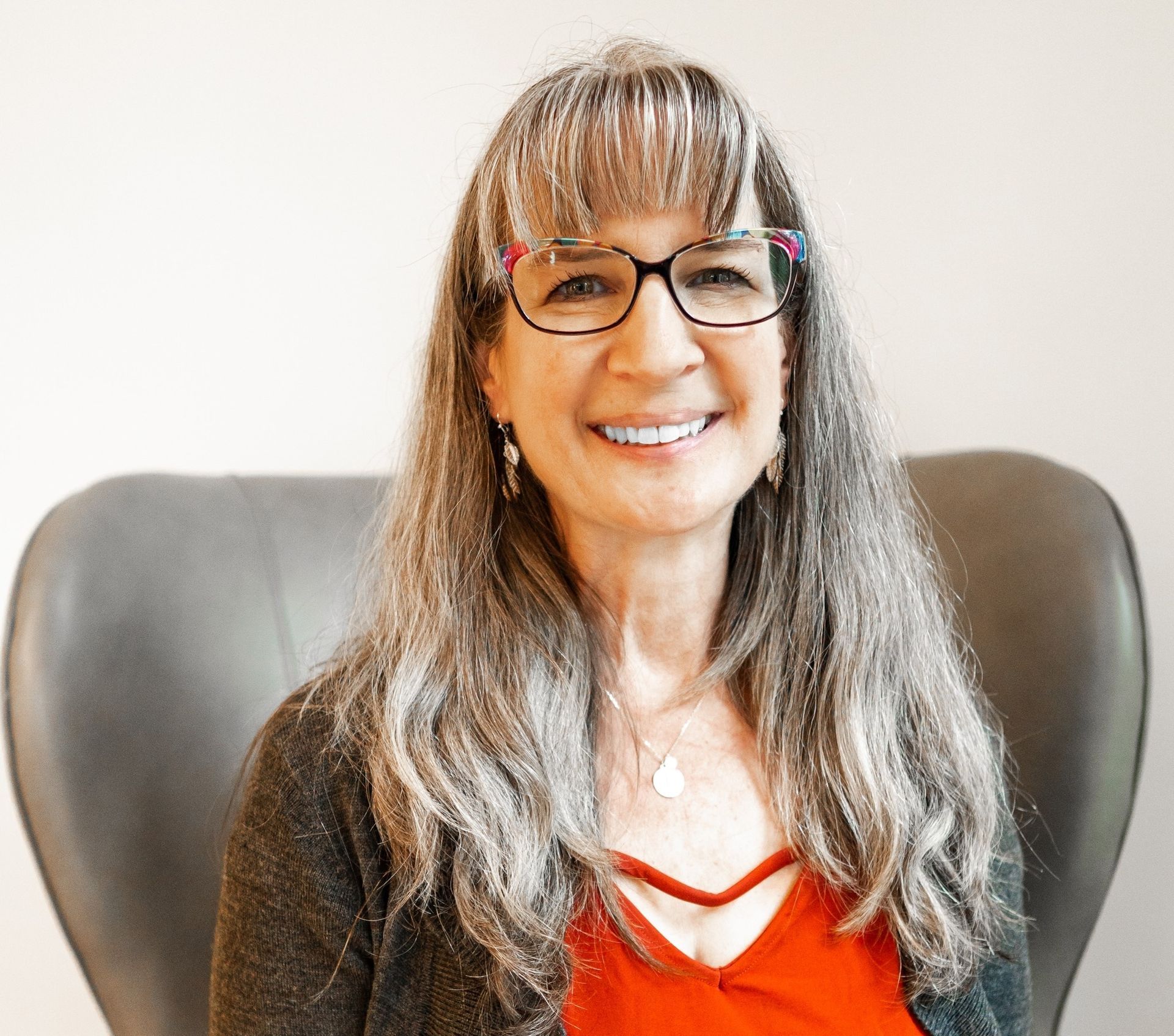 A woman wearing glasses and a red shirt is sitting in a chair and smiling.