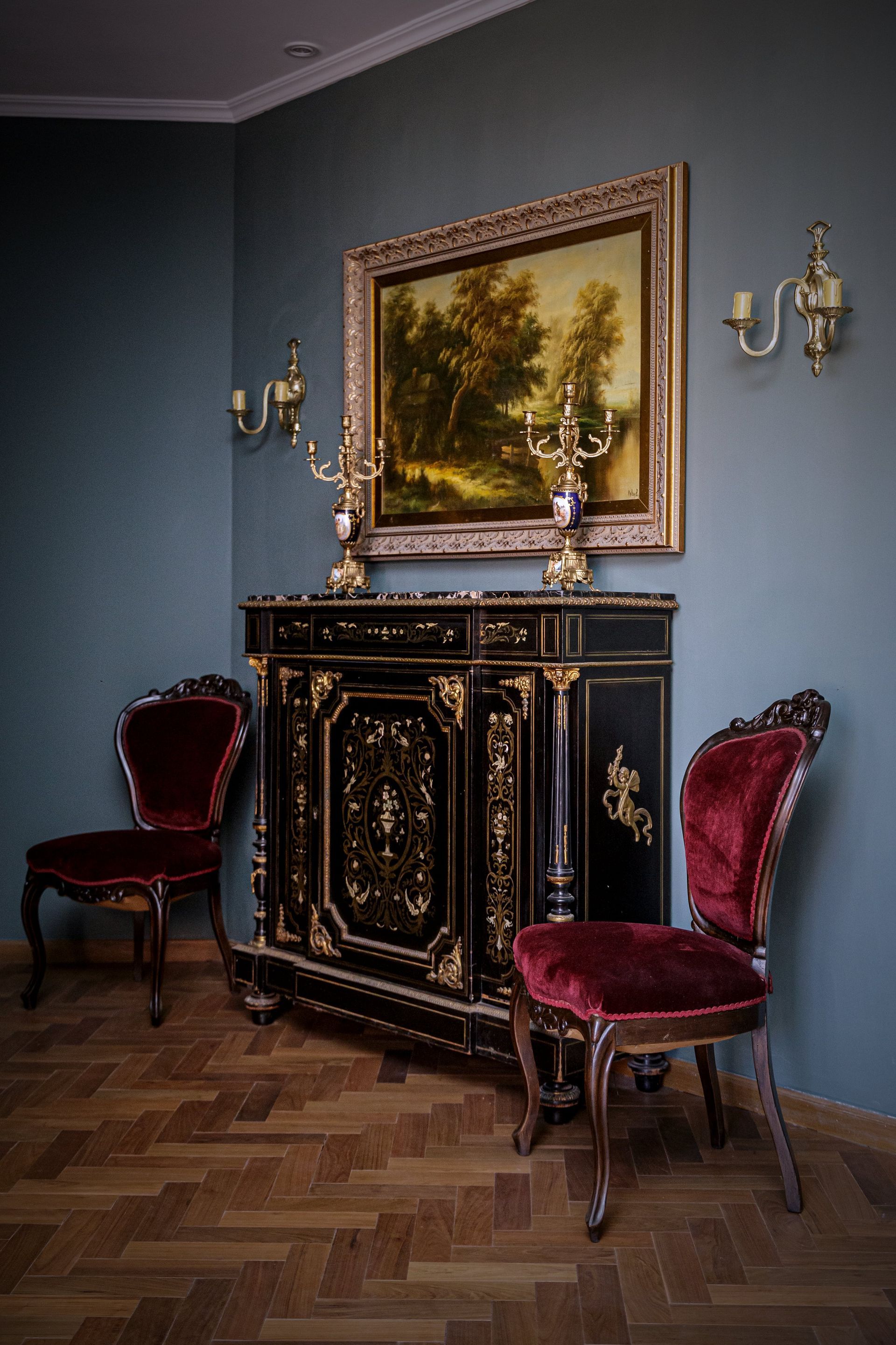 Una pintura de paisaje enmarcada, dos sillas de terciopelo burdeos y apliques.
