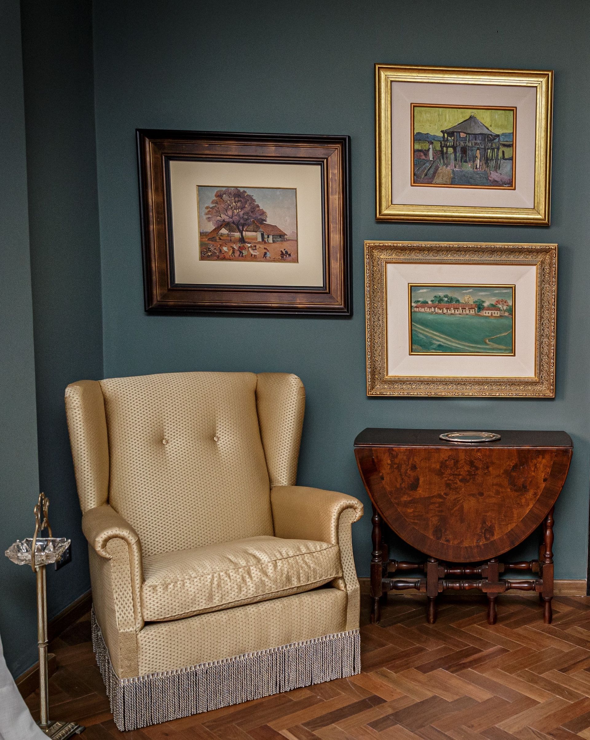 Sillón orejero, cuadro enmarcado y mesa antigua sobre una pared verde azulado. Suelo de madera.