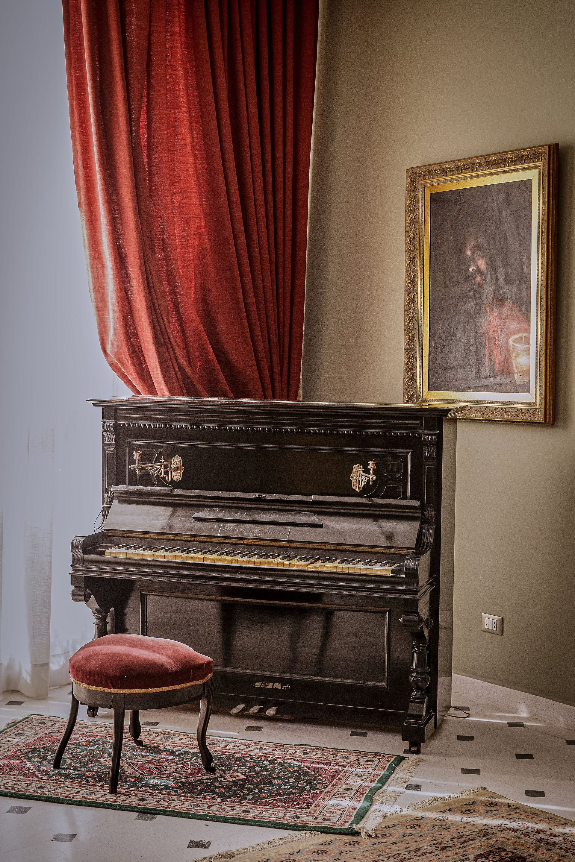 Piano vertical negro con taburete de terciopelo rojo, cortinas rojas y un cuadro enmarcado sobre una pared color canela.