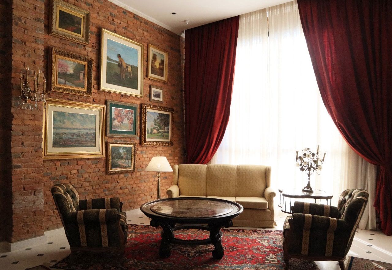 Acogedora sala de estar con pared de ladrillo, arte enmarcado, cortinas rojas, sofá pequeño, sillones y mesa redonda.