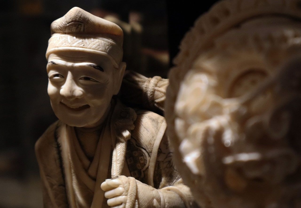 Figura de marfil: hombre sonriente con sombrero y túnica, sosteniendo un bastón. Otra figura se ve borrosa al fondo.