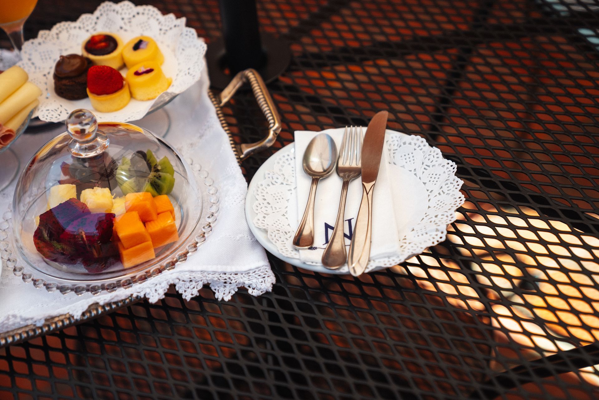 Un servicio de mesa con una fuente de fruta bajo una cúpula de cristal y un plato con cubiertos sobre una mesa de rejilla de metal negro.