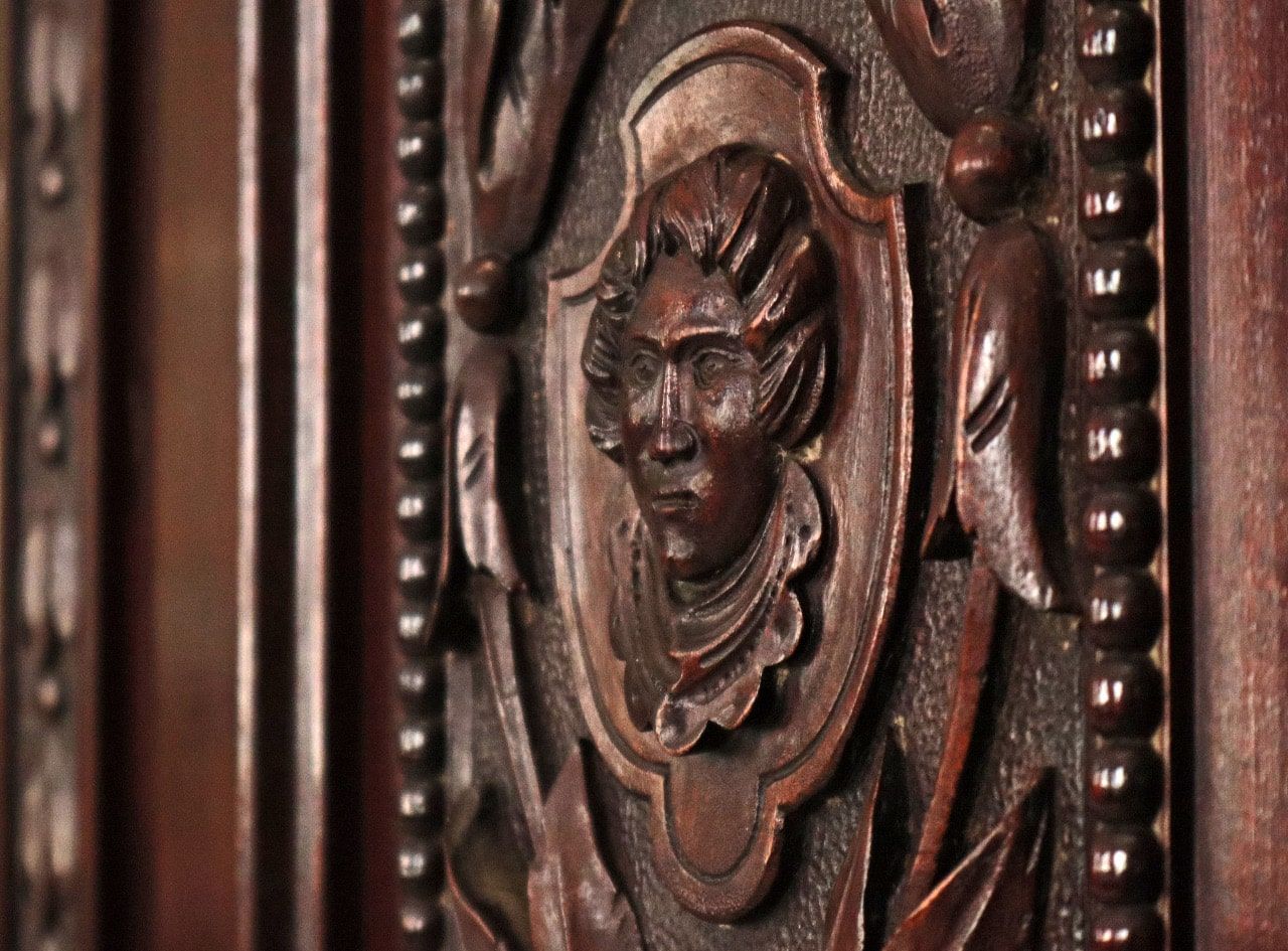 Primer plano de un panel de madera tallada; un rostro dentro de un marco ornamentado, rodeado de hojas.