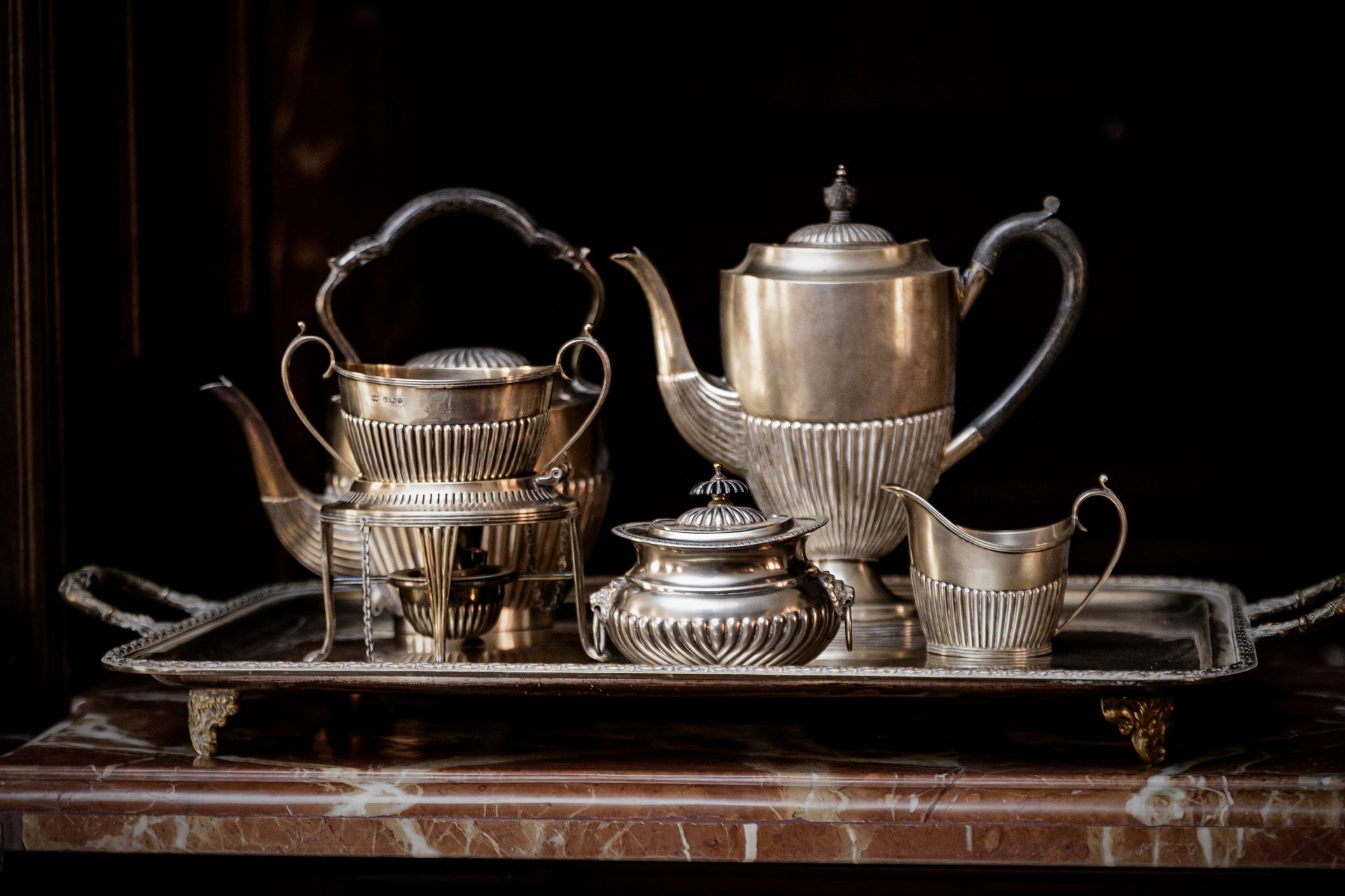 Juego de té de plata en bandeja, con tetera, azucarero, cremera y calentador. Fondo oscuro.
