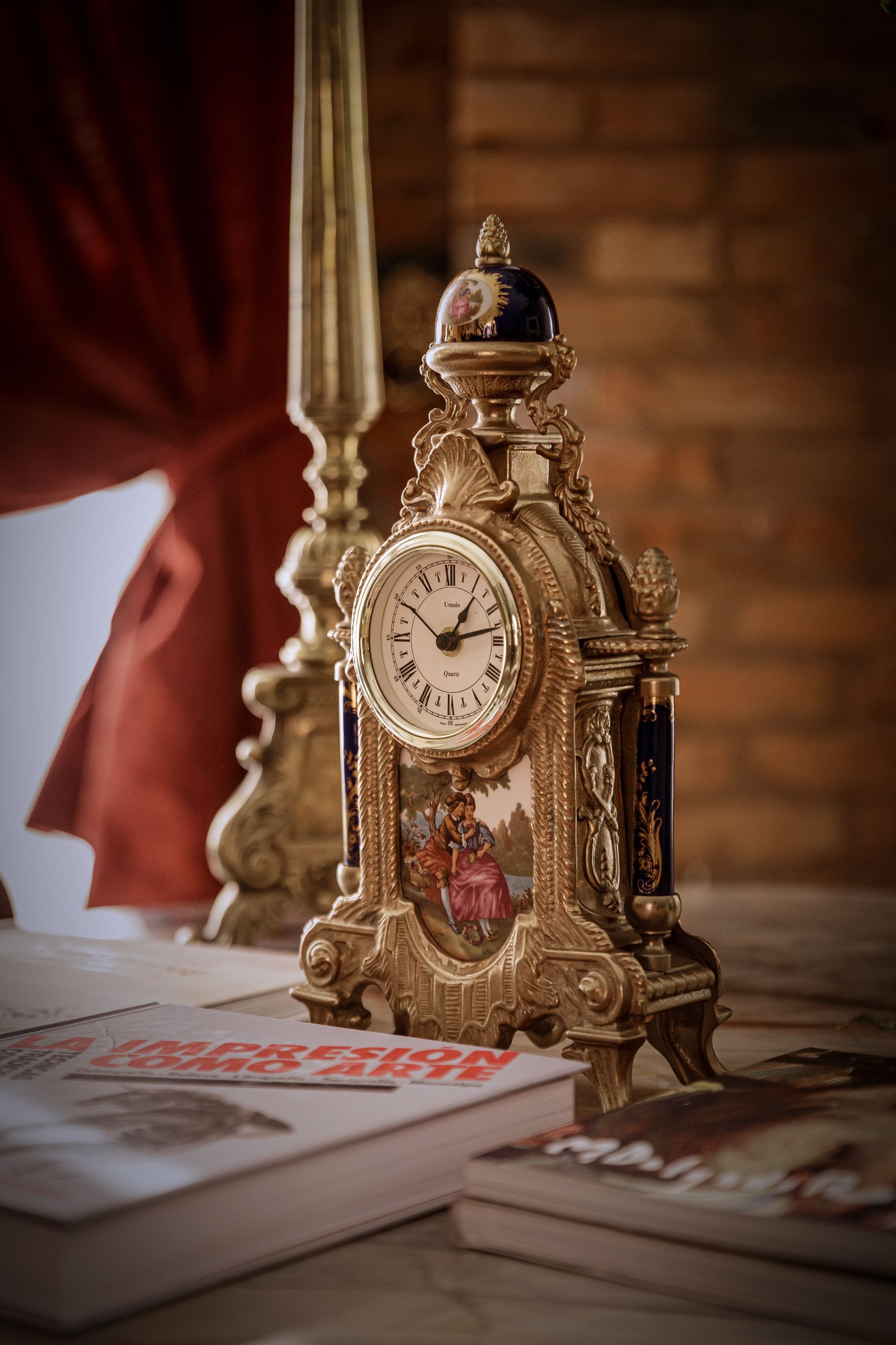 Reloj dorado adornado sobre una mesa con libros, candelabro y cortina roja.