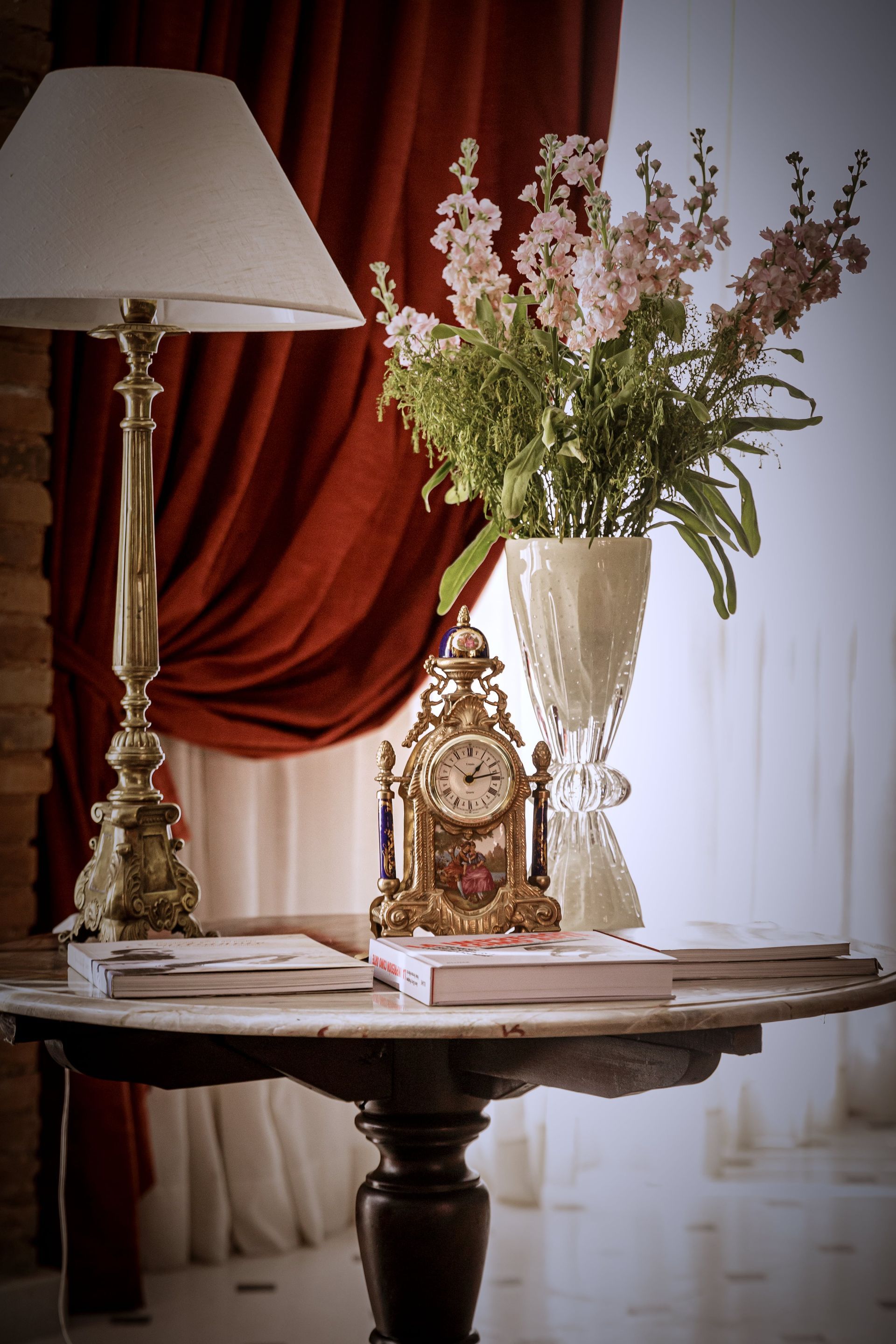 Una mesa con una lámpara, un jarrón de flores, un reloj decorativo y libros frente a una cortina roja.