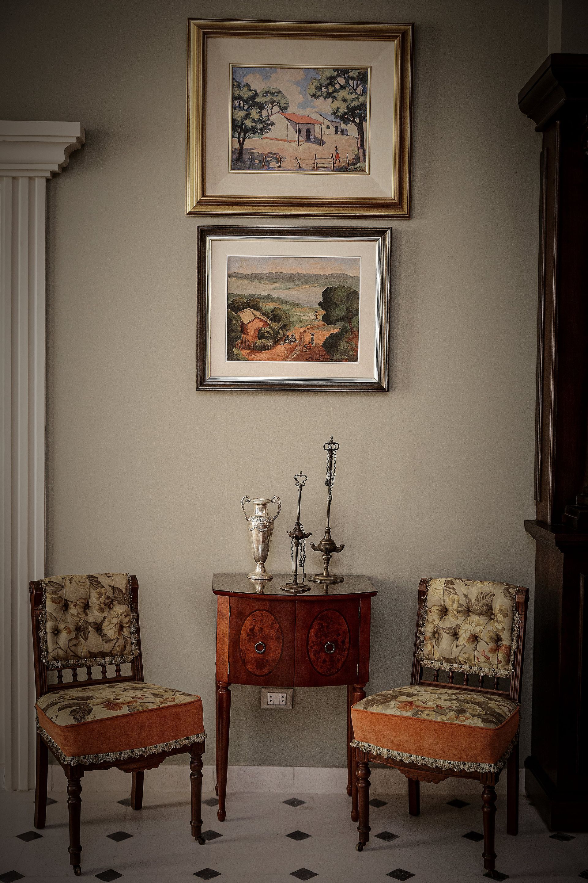 Dos sillas flanquean un armario debajo de pinturas de paisajes en una habitación bien iluminada.