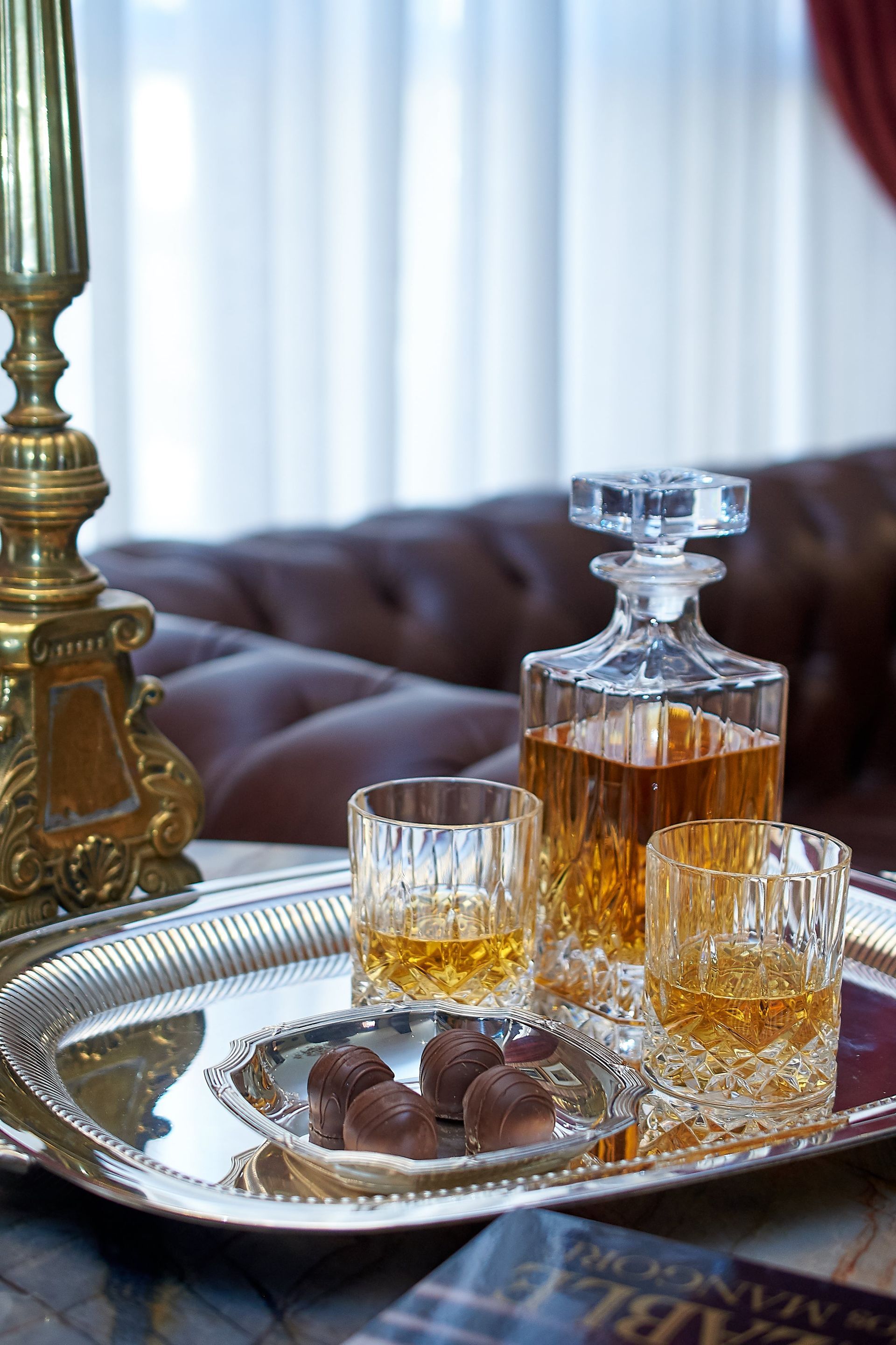 Decantador de whisky y vasos en una bandeja de plata con bombones, junto a un sofá de cuero y una lámpara.
