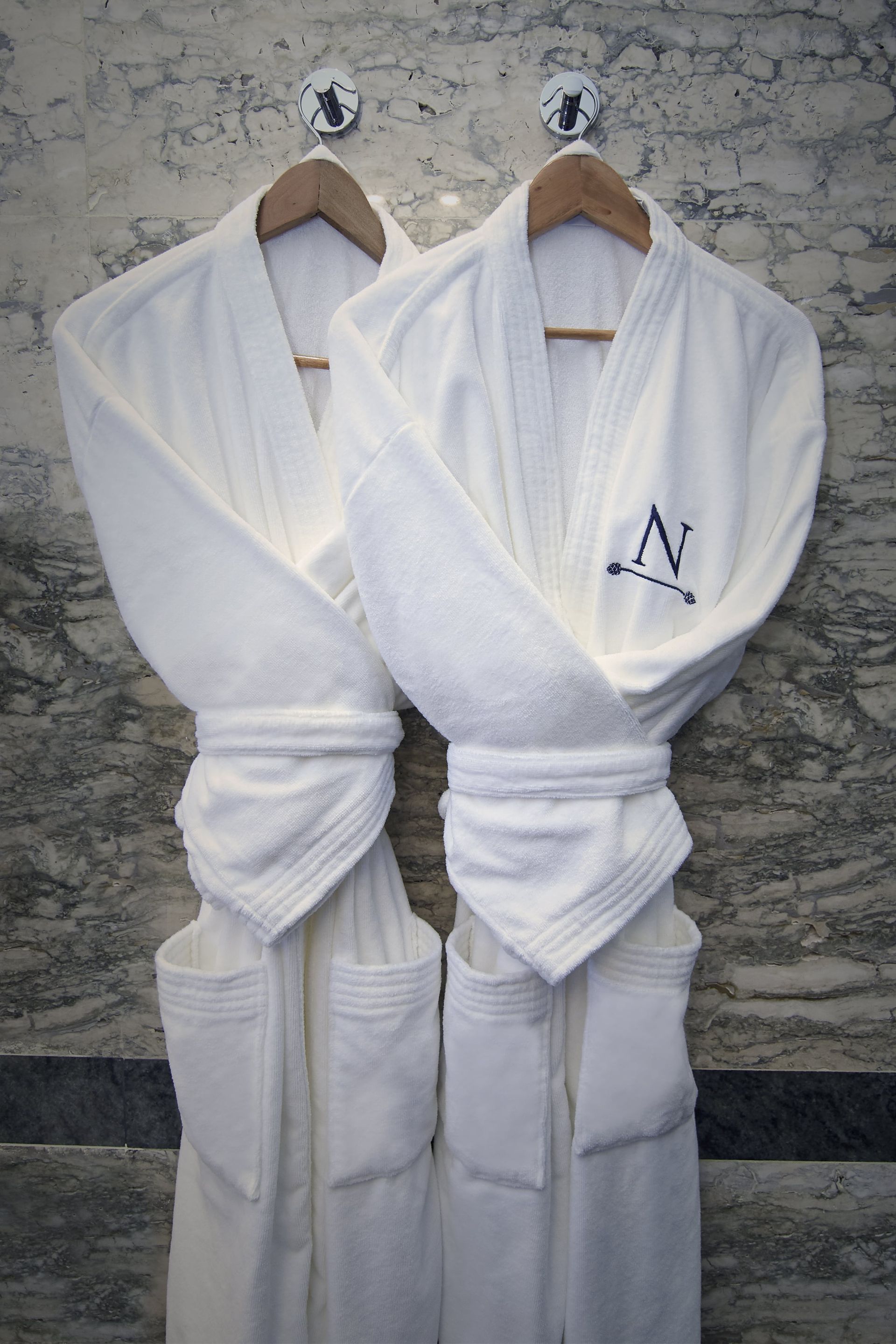 Dos albornoces blancos colgados en perchas de madera. Uno tiene la letra 