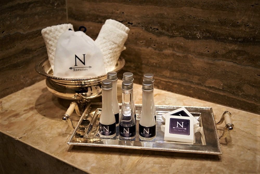 Baño con bandeja de plata que contiene artículos de tocador y toallas en una cesta de plata, contra una pared de mármol.