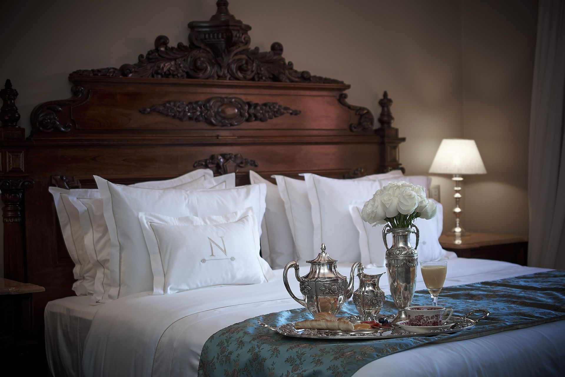 Elegante dormitorio con cabecera de madera ornamentada, ropa de cama blanca y juego de té plateado sobre una bandeja de cama.