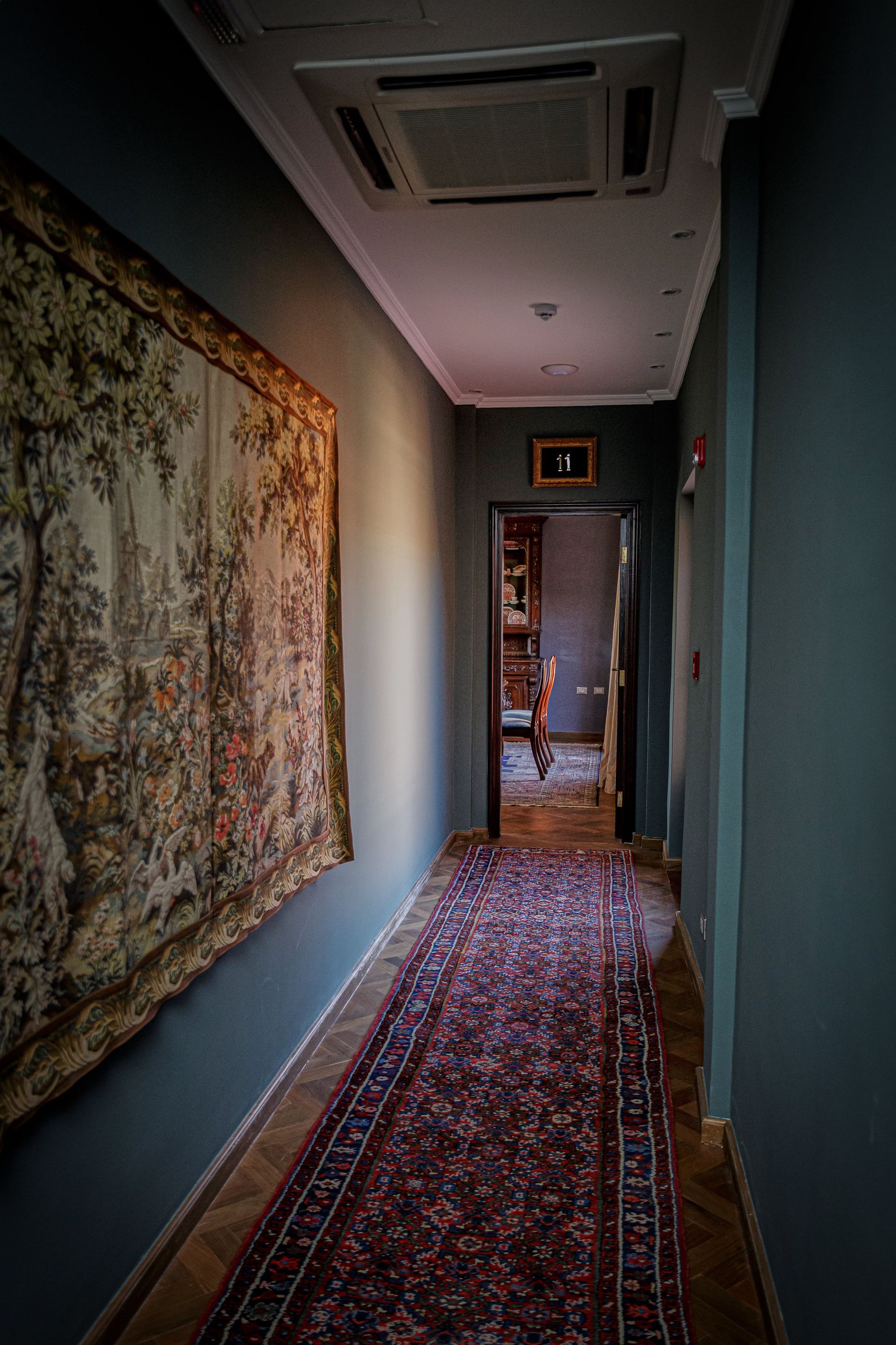 Pasillo largo con paredes de color verde azulado oscuro, alfombra floral, tapiz y puerta que conduce a una habitación.