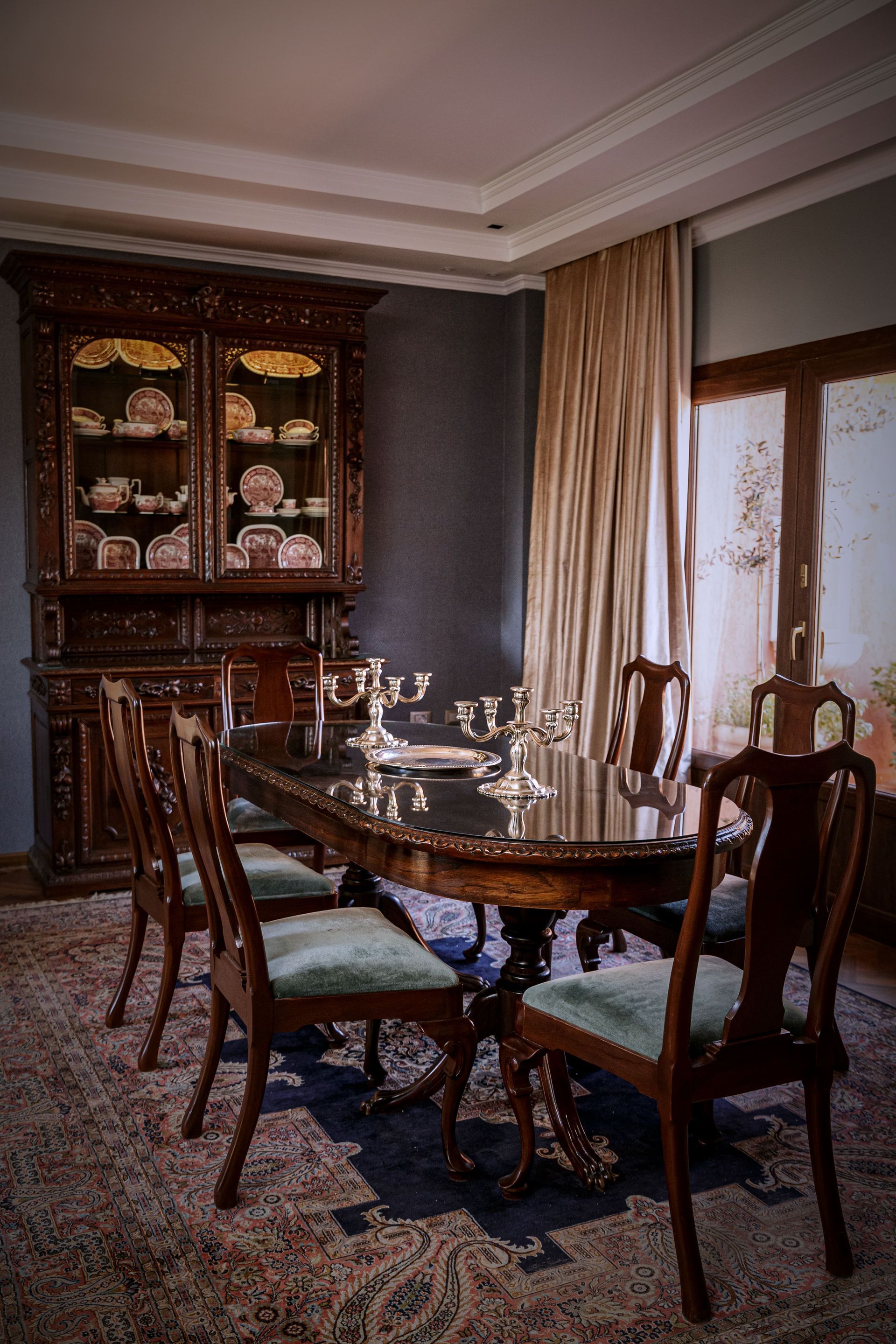 Comedor con muebles de madera oscura: vitrina, mesa puesta con plata y sillas sobre alfombra estampada.