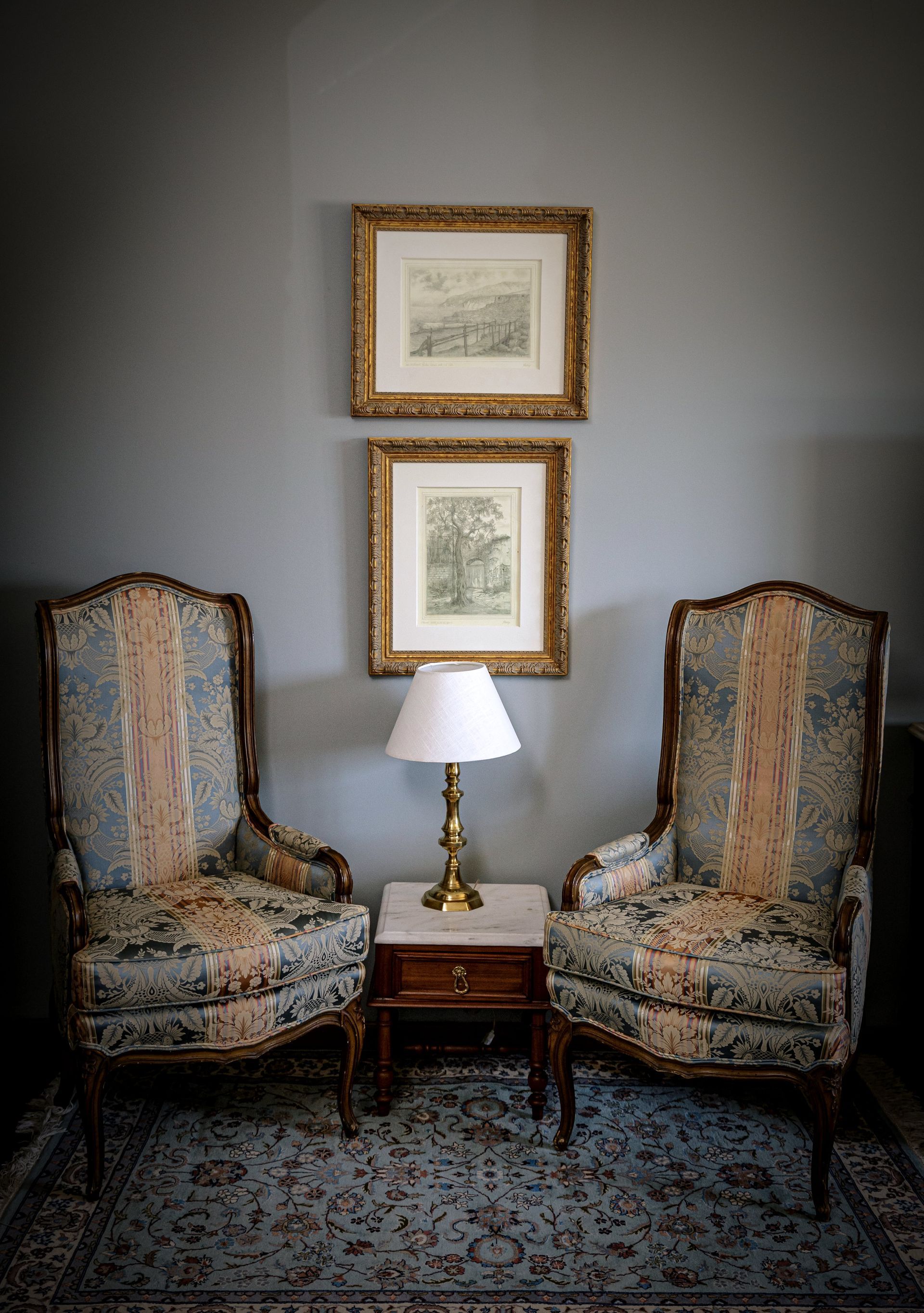 Dos sillones ornamentados flanquean una pequeña mesa con lámpara y obras de arte en una pared gris.