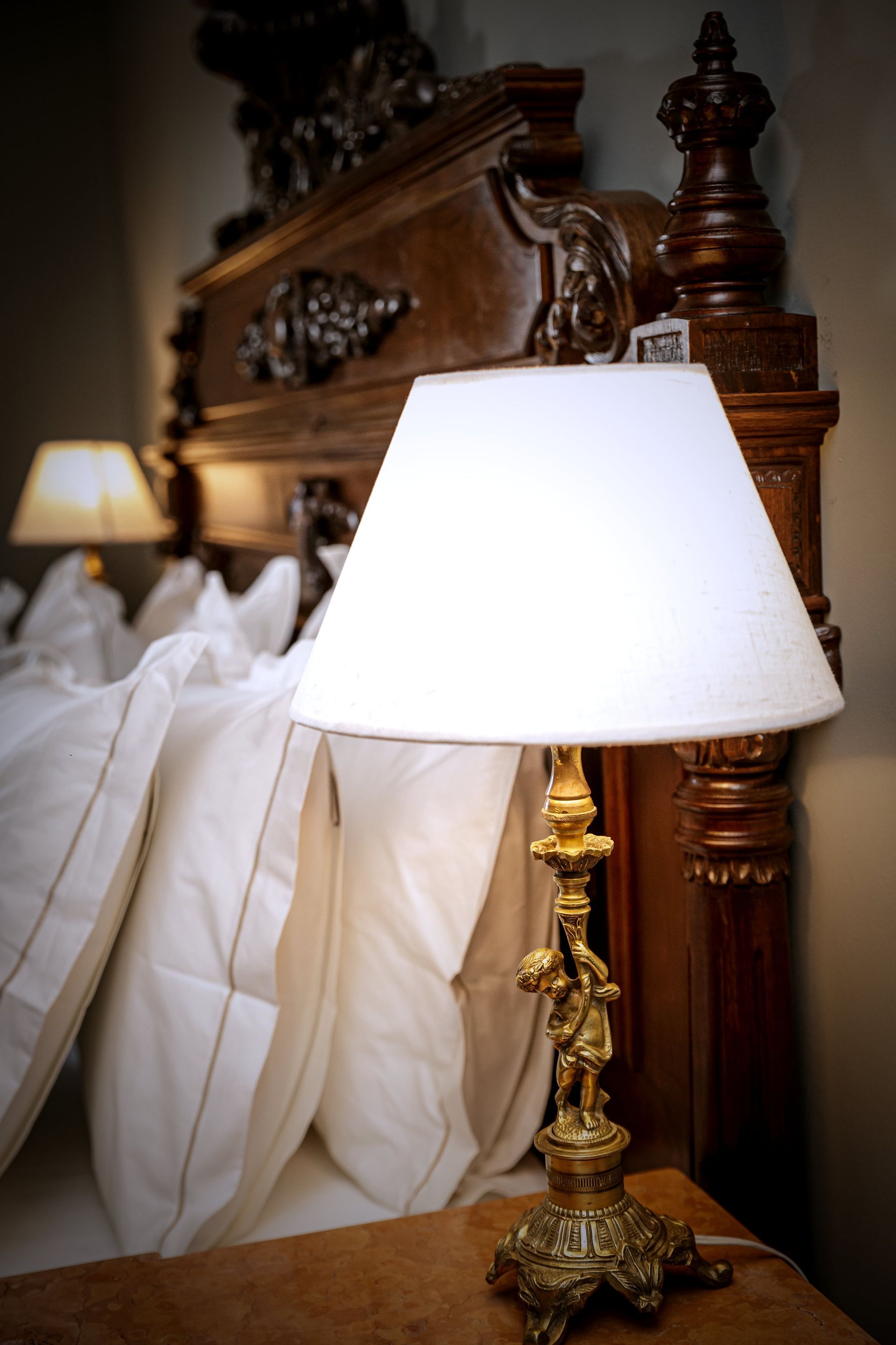 Lámpara de latón ornamentada sobre una mesita de noche junto a una cama con sábanas blancas y un cabecero de madera tallada.