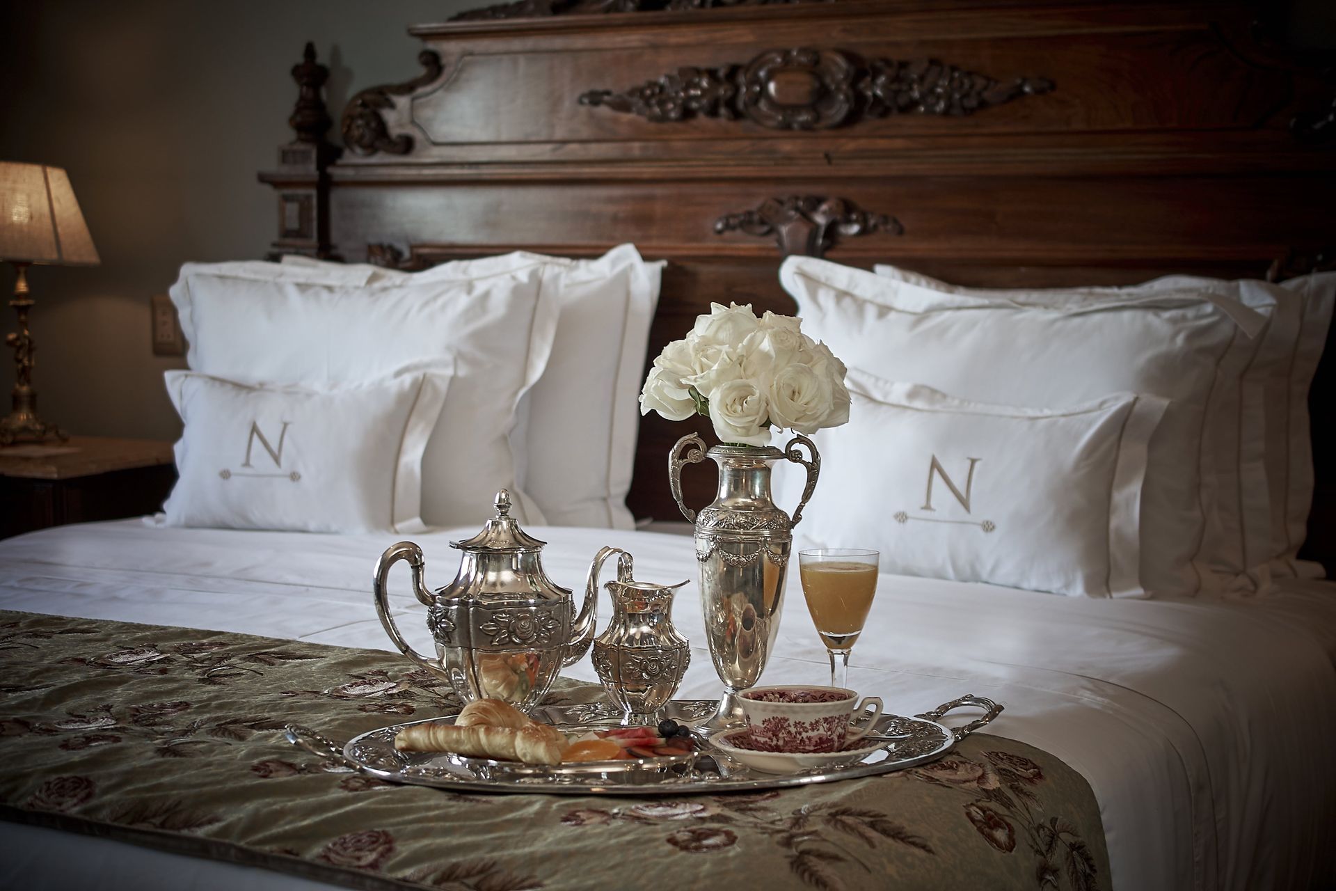 Bandeja de desayuno sobre la cama con juego de plata y flores en un lujoso dormitorio.