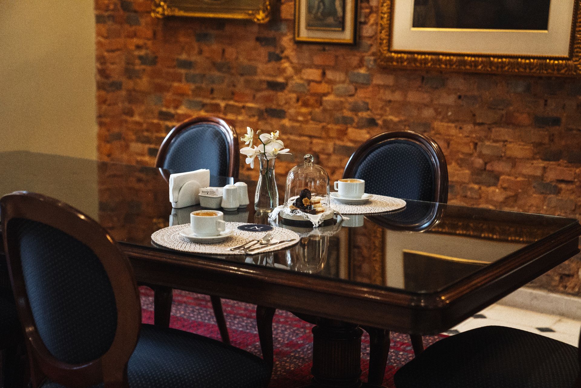 Una mesa de comedor con tablero de cristal, sillas, tazas de café y un jarrón, dispuesta contra una pared de ladrillo rústico de tonos cálidos.