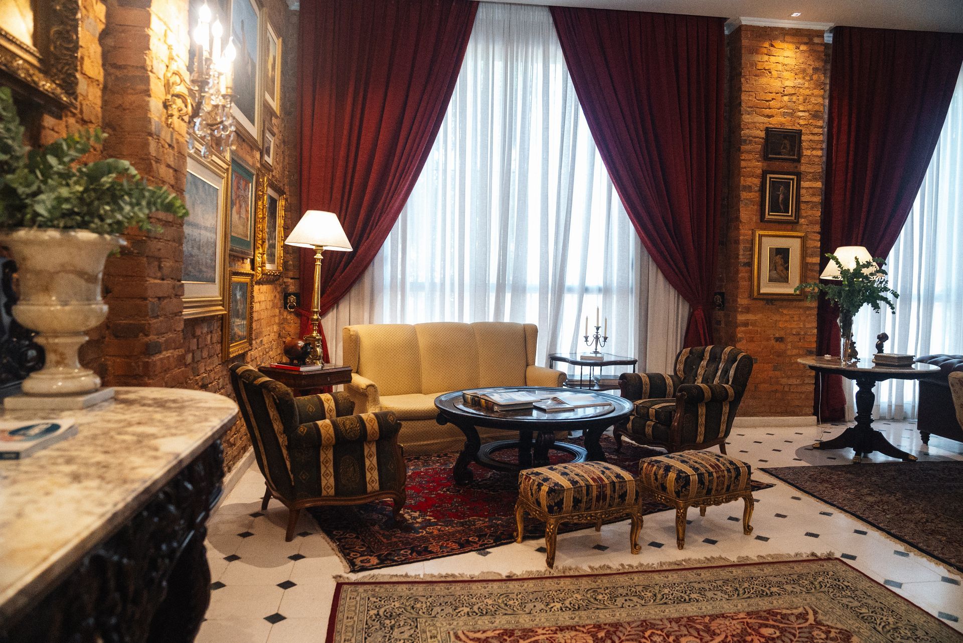 Una habitación lujosa y con iluminación tenue, con paredes de ladrillo visto, cortinas ornamentadas, sillones estampados, un sofá y alfombras sobre un suelo de mármol.