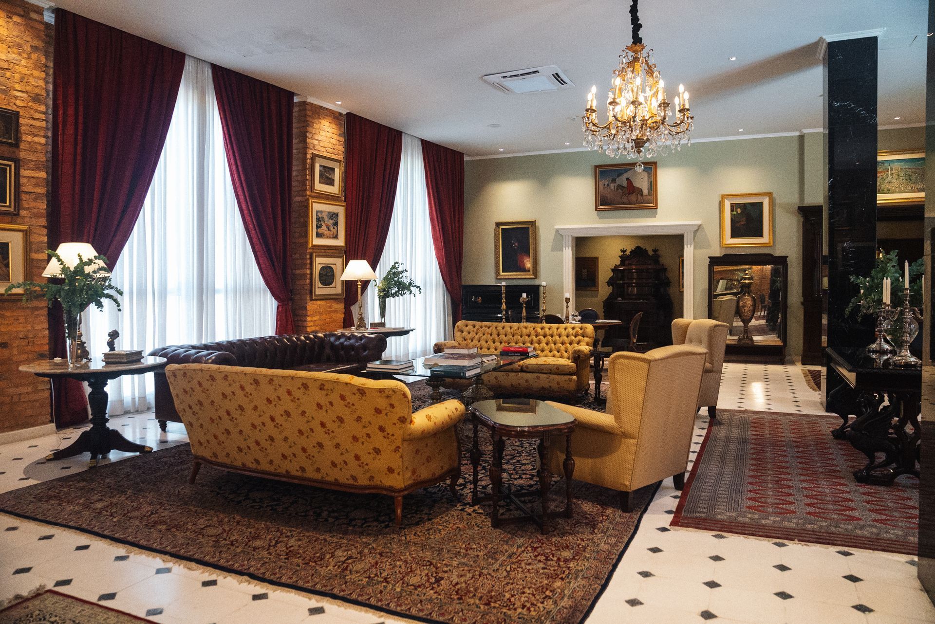 Un elegante salón luminoso con sofás amarillos, lámparas de araña antiguas, cortinas granates y cuadros enmarcados sobre suelos de baldosas.