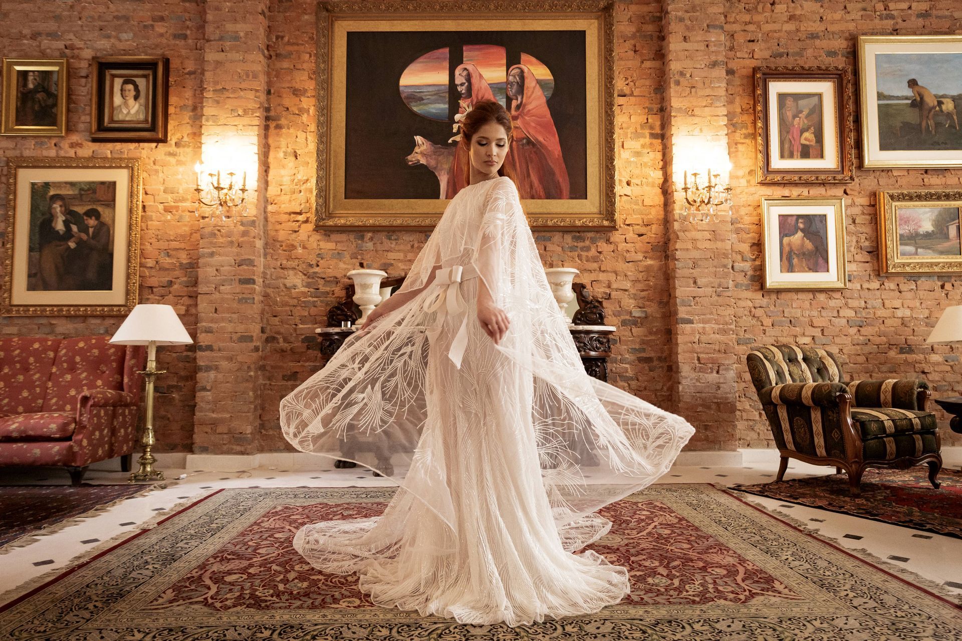 Mujer con vestido blanco dando vueltas en una habitación adornada con pinturas y una pared de ladrillos rojos.