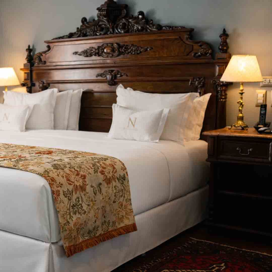 Elegante dormitorio con cabecera de madera ornamentada, ropa de cama blanca y juego de té plateado sobre una bandeja de cama.