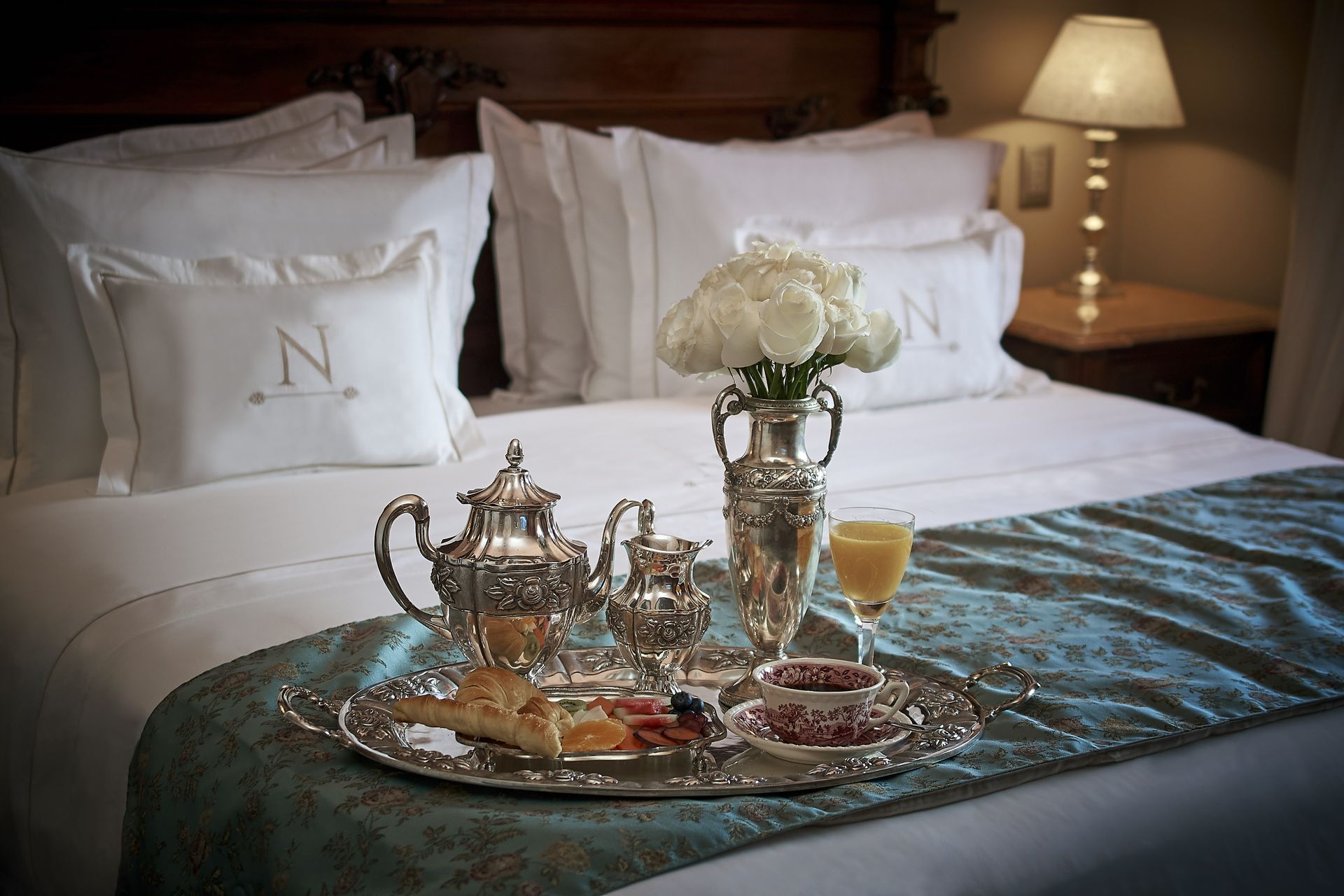 Bandeja de desayuno sobre una cama con sábanas blancas; juego de té de plata, flores, jugo.