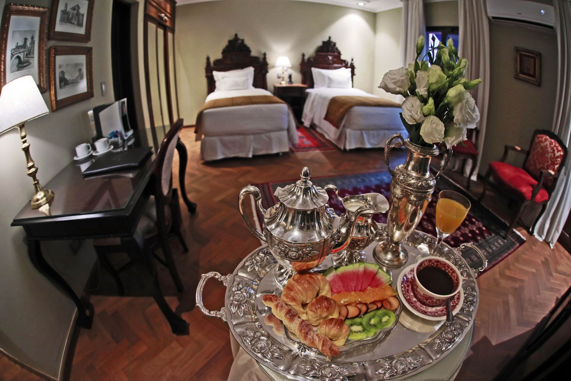 Habitación de hotel con dos camas individuales, bandeja de desayuno, escritorio y arreglo floral.
