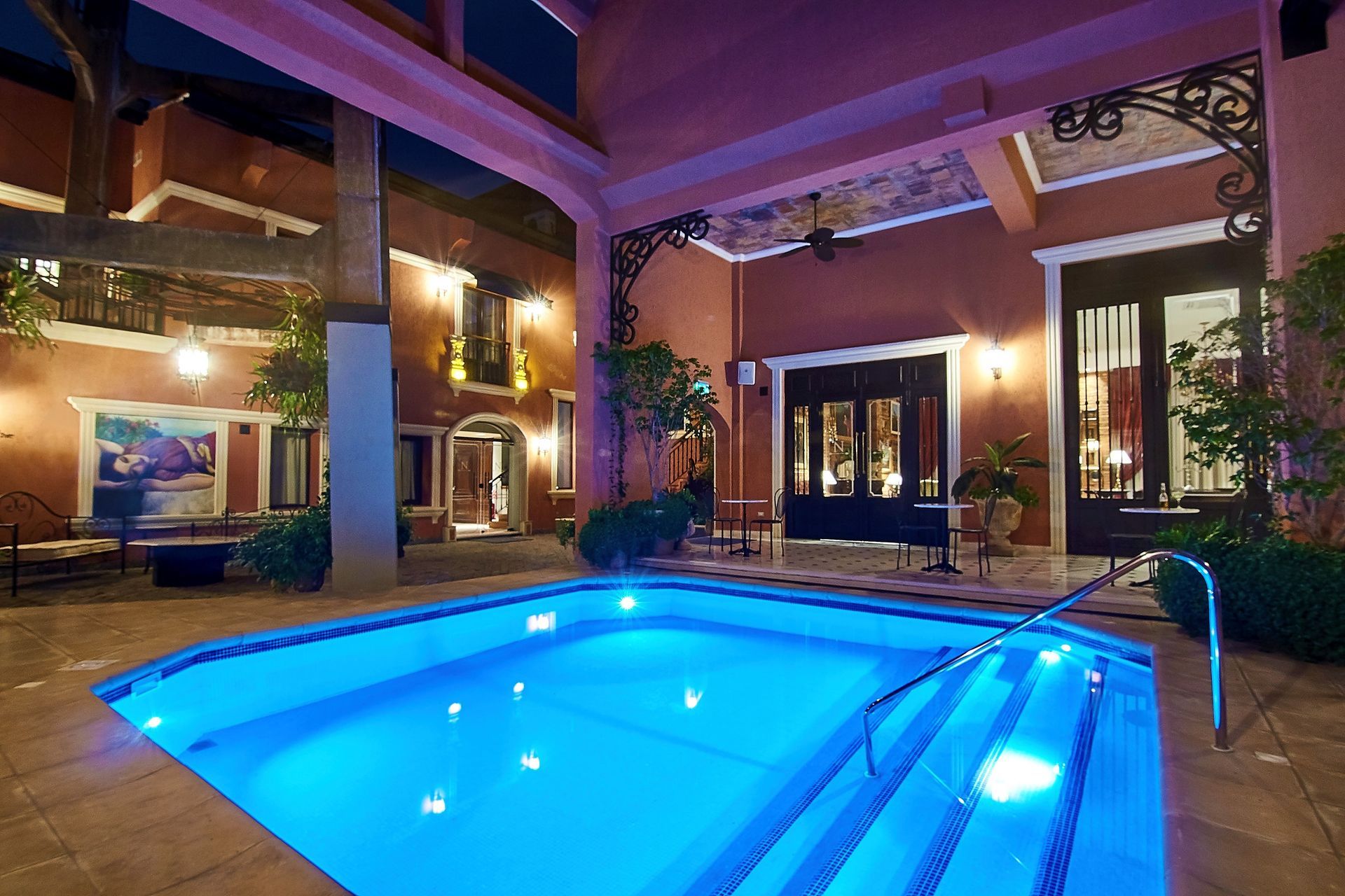 Pequeña piscina iluminada en un patio rodeado de edificios con detalles arquitectónicos ornamentados, por la noche.