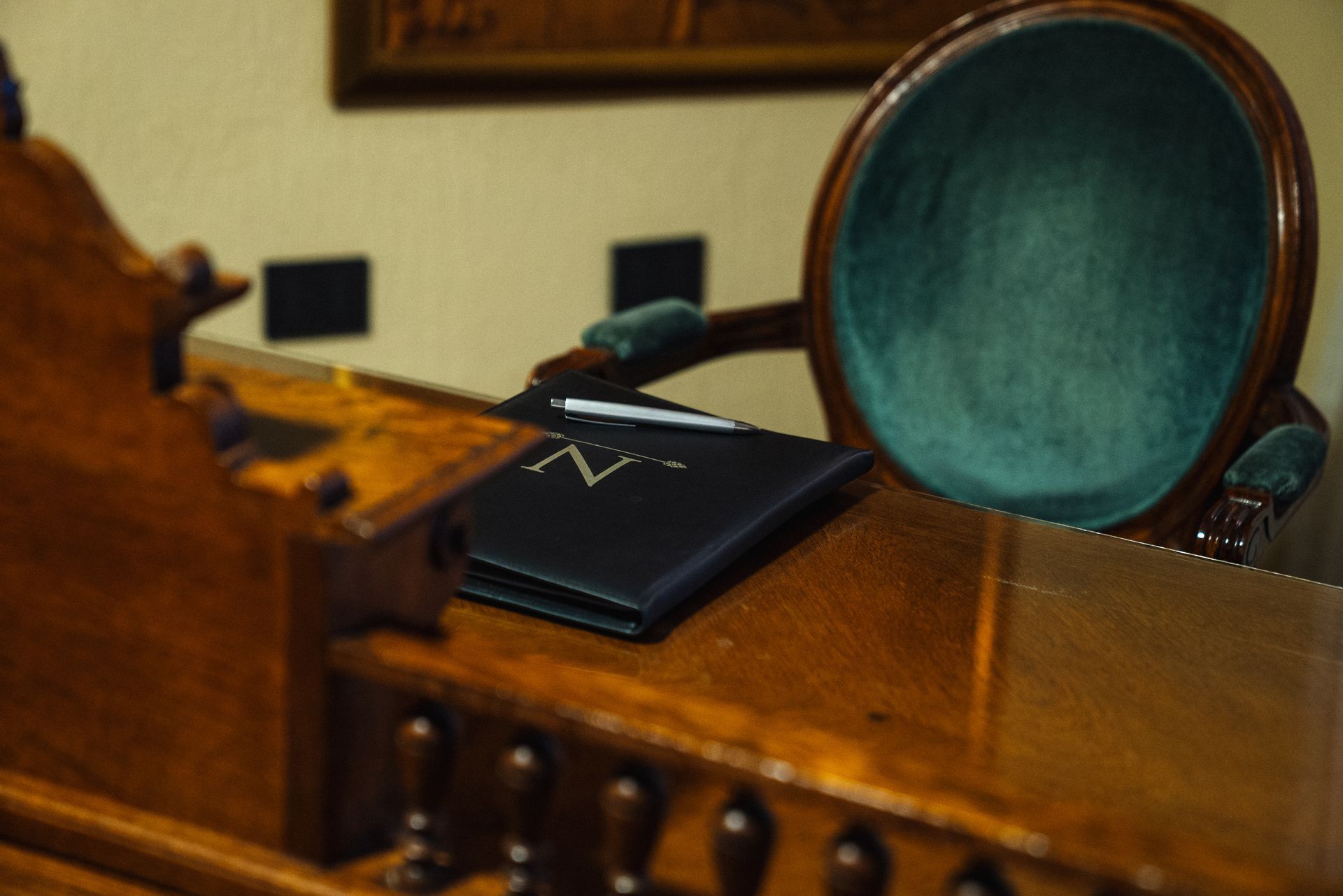 Un cuaderno azul oscuro con un logotipo metálico reposa sobre un escritorio de madera junto a una silla de terciopelo verde en una oficina con poca luz.