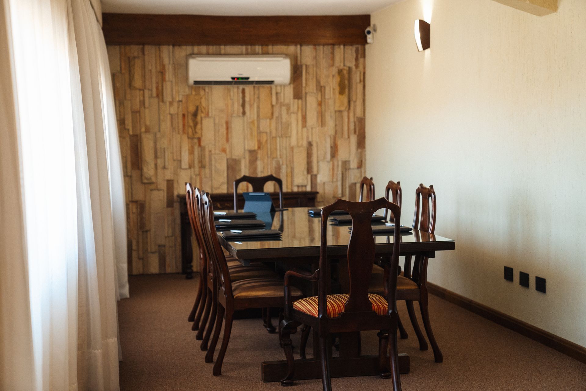Una sala de reuniones con una mesa y sillas de madera oscura, y un panel de pared de madera clara con textura, todo ello bajo un aire acondicionado.