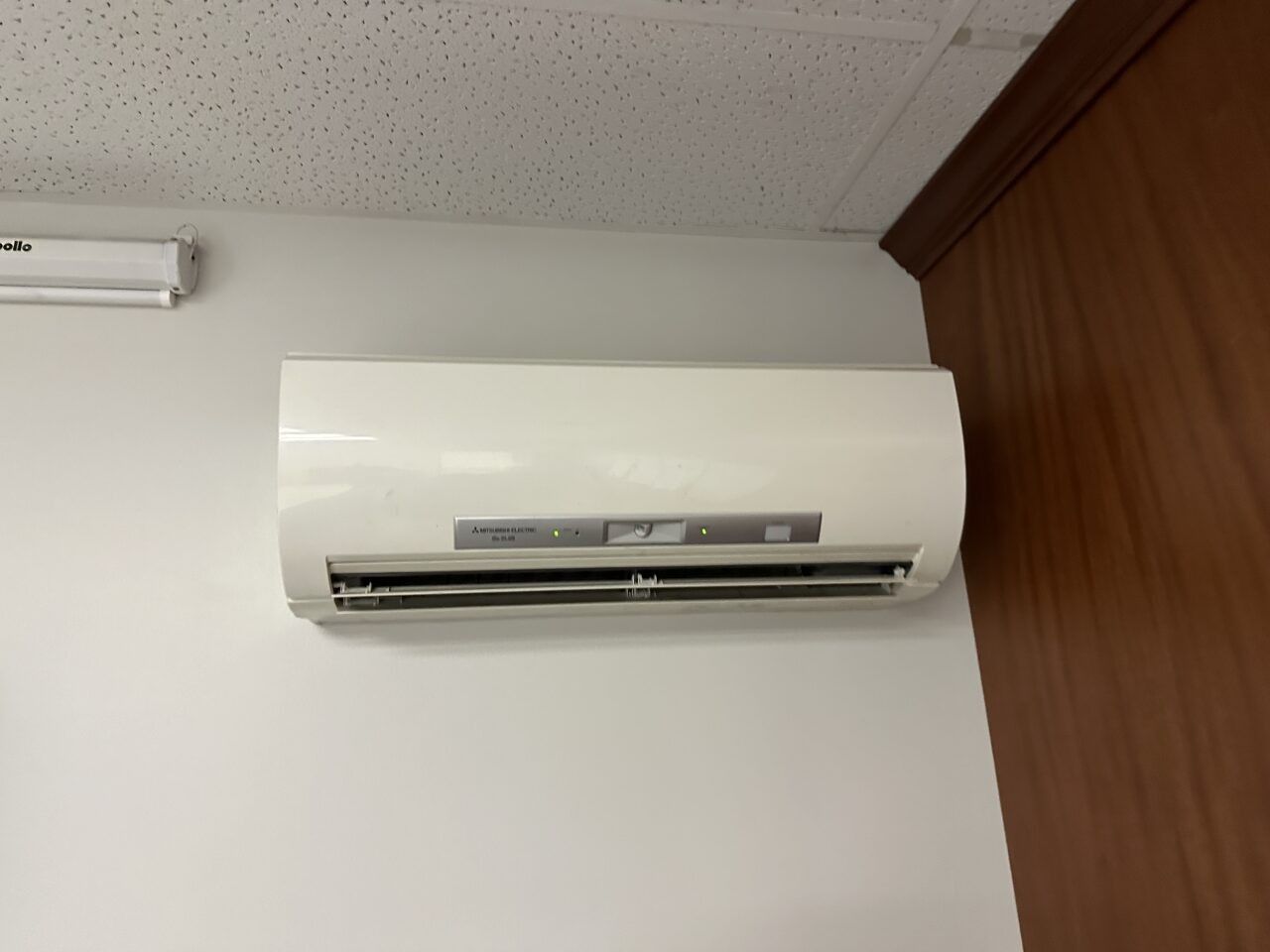 A white wall-mounted air conditioning unit installed high on a wall next to a wooden panel.