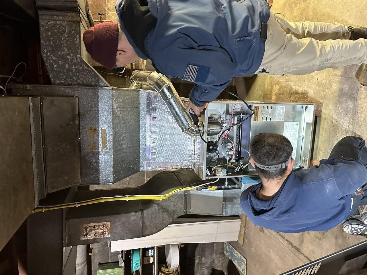 Two service technicians in blue uniforms inspect an open furnace unit in a basement.