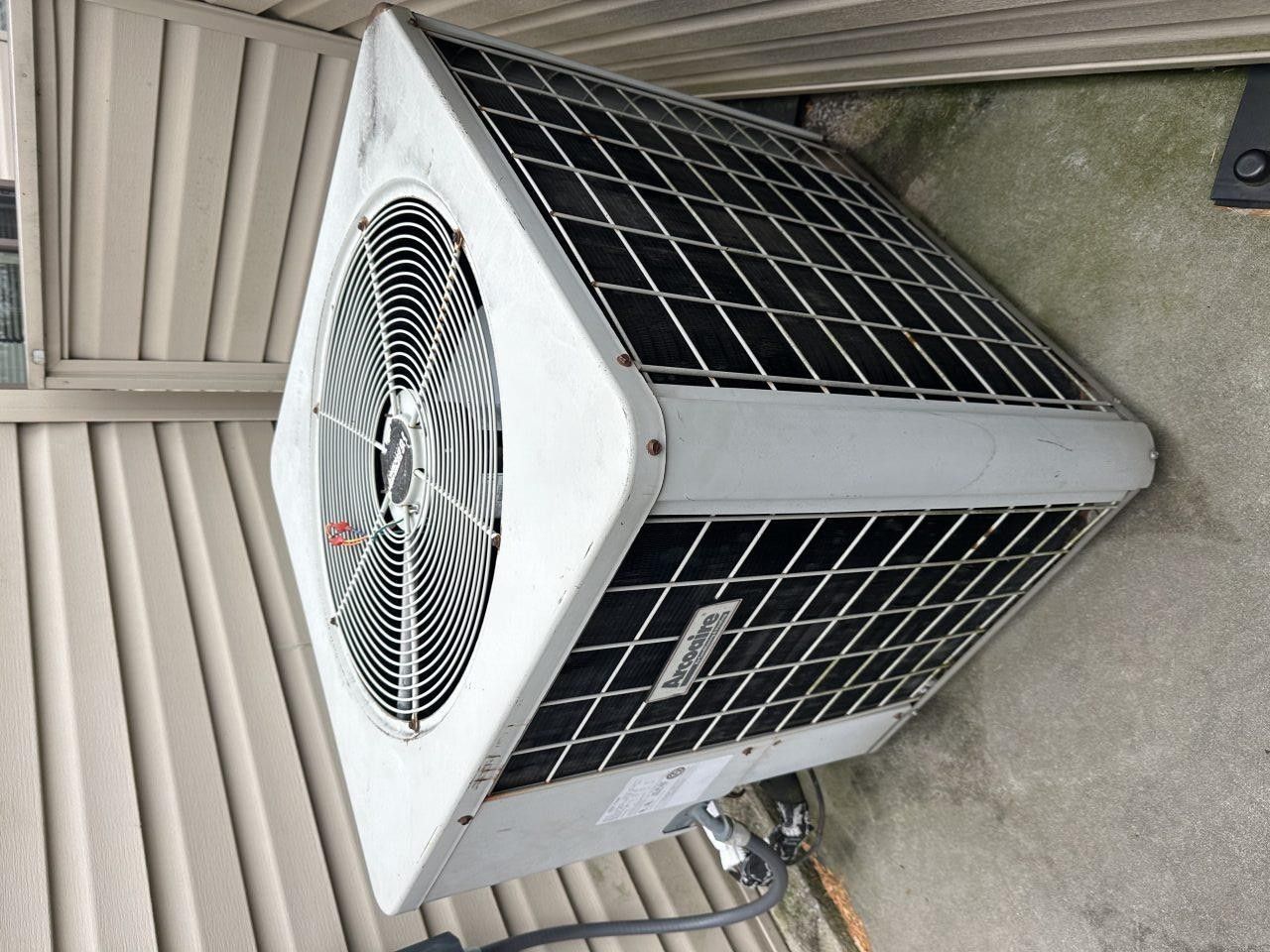 A white outdoor air conditioning condenser unit sits on a concrete patio against a beige siding wall.