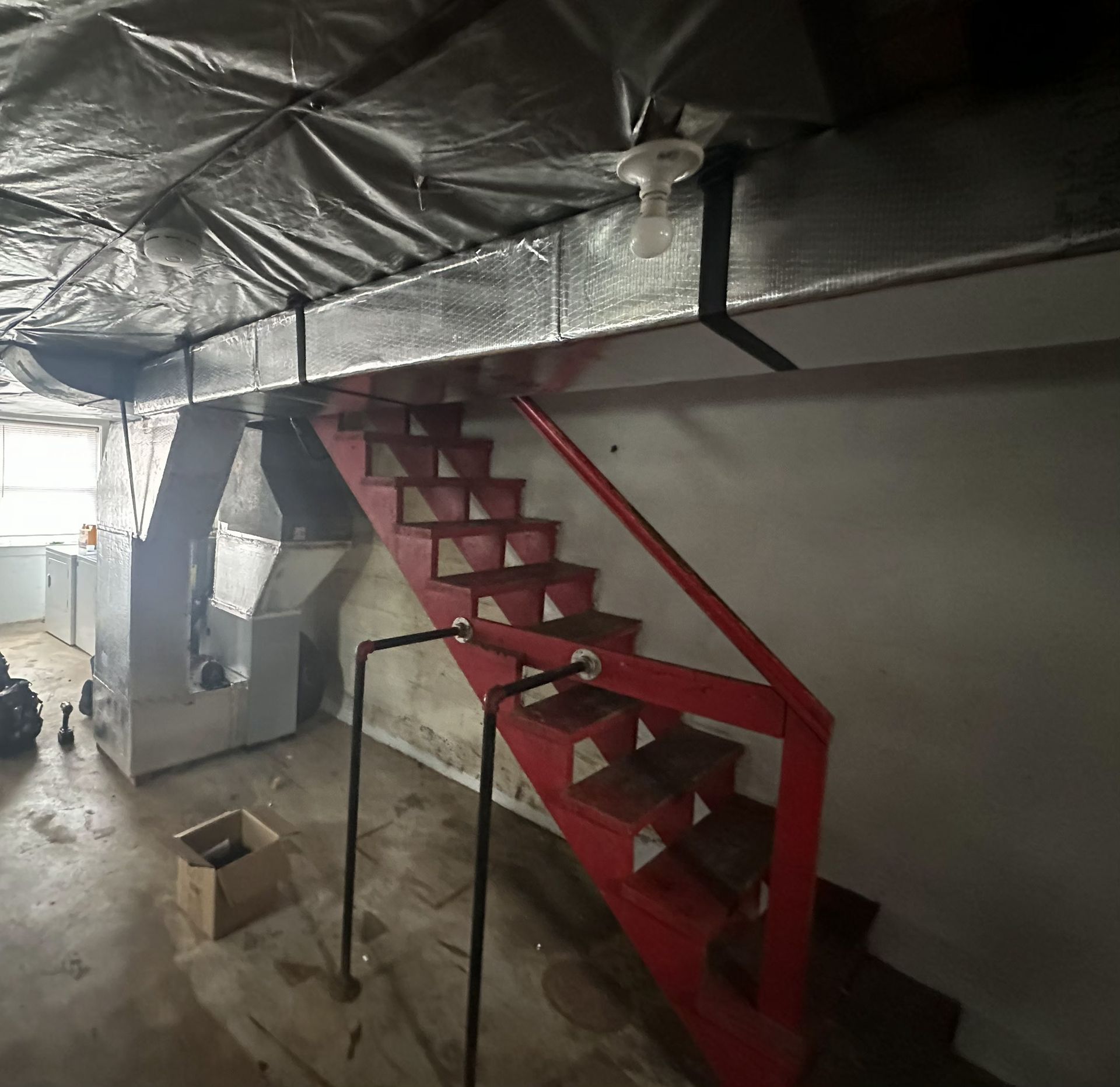 A red metal staircase leads upward in a basement with concrete floors and foil-insulated ductwork on the ceiling.