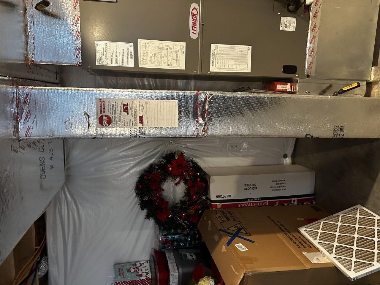 An HVAC furnace unit and silver ductwork inside a cluttered storage room with boxes, a holiday wreath, and a spare filter.