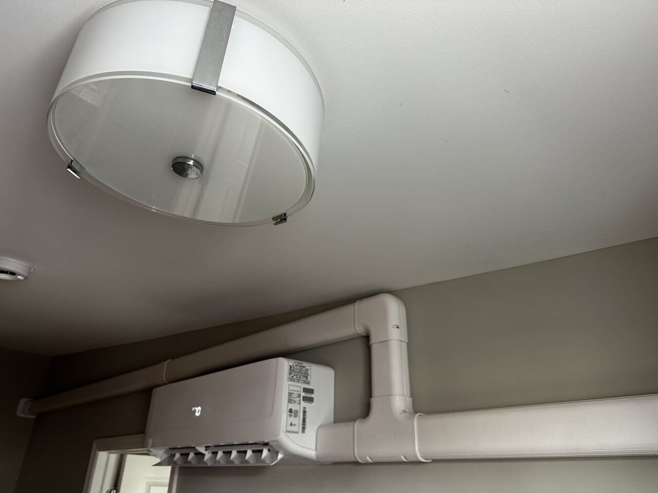 A white indoor wall-mounted air conditioning unit with connected PVC trunking mounted on a wall below a ceiling light.