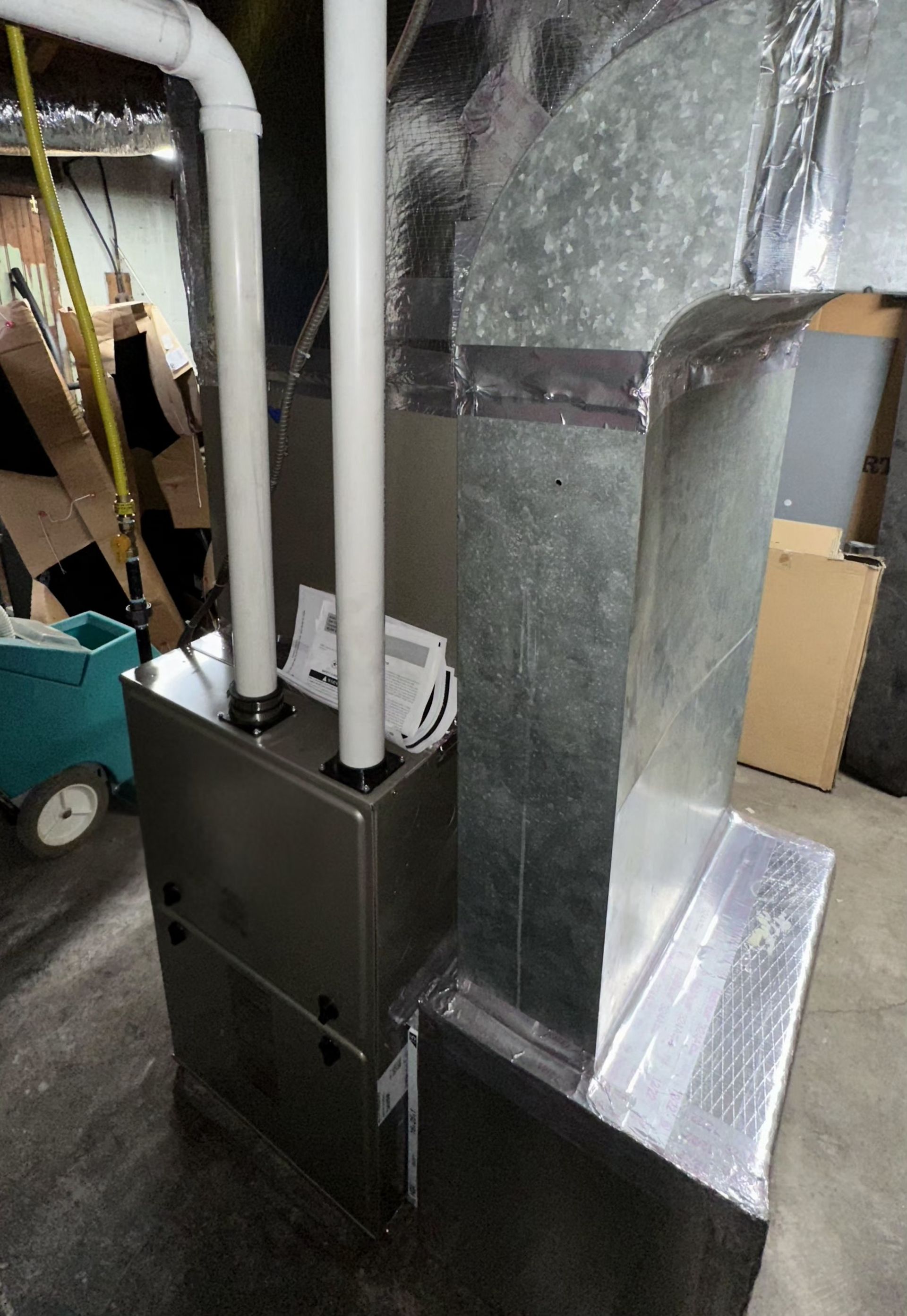 A furnace with two white PVC exhaust pipes stands next to a large, rectangular metal duct in a residential basement.