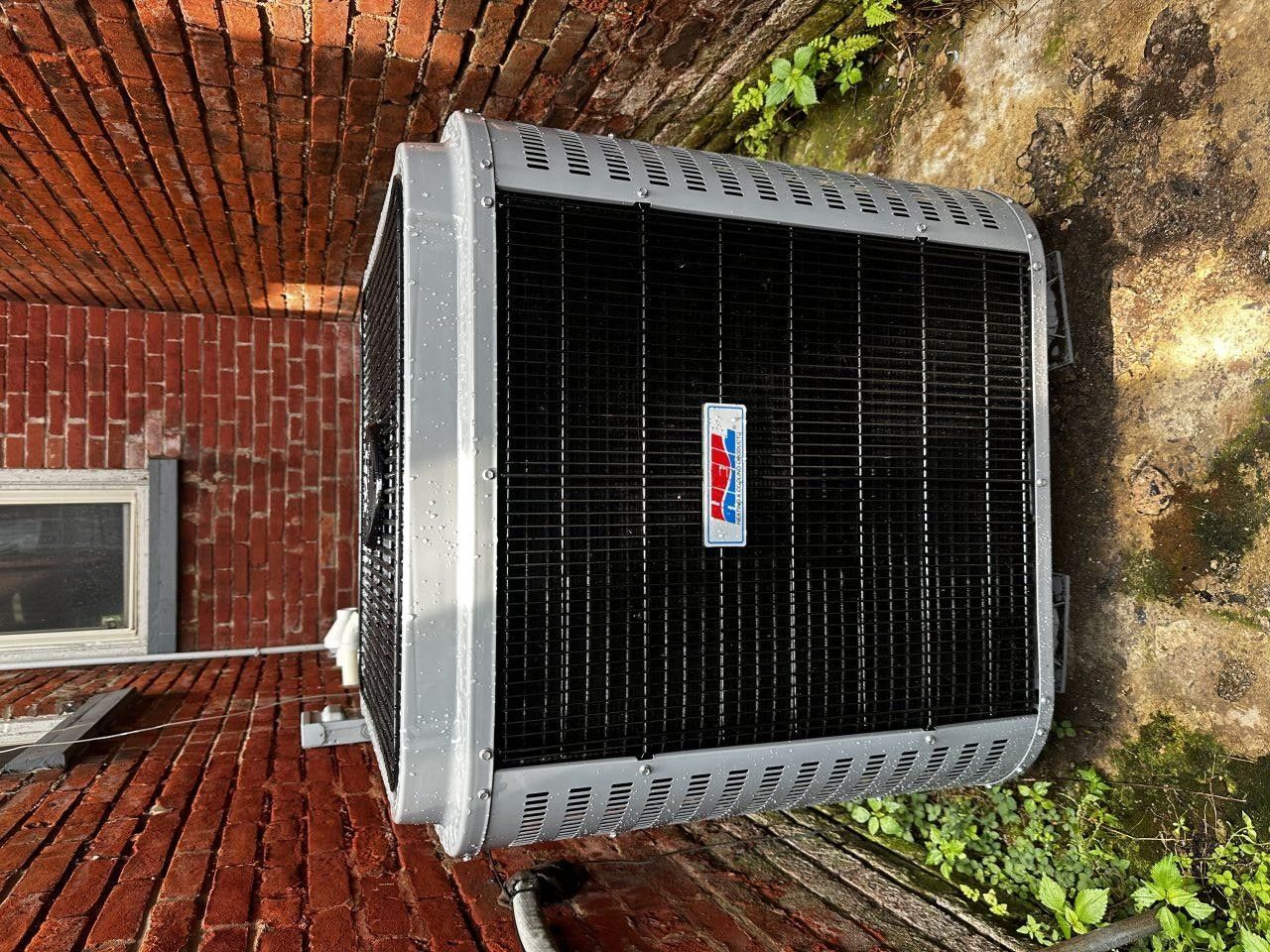 An American Standard brand outdoor air conditioning unit sits on the ground next to a red brick wall.