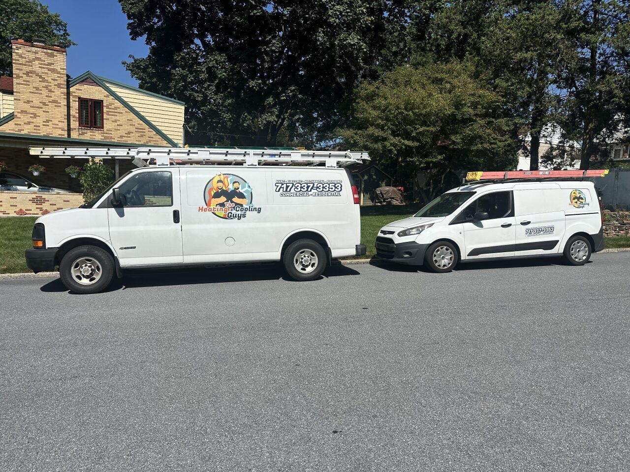 Two white service vans with ladders on their roofs parked on a paved lot in front of a house.