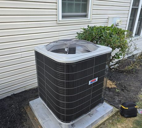 A gray residential air conditioning unit sits on a concrete pad outside a house with white siding and a green bush.