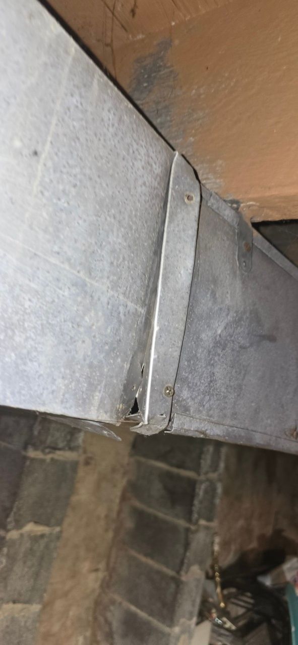 A damaged section of metallic HVAC ductwork showing a disconnected joint hanging below an unfinished ceiling.