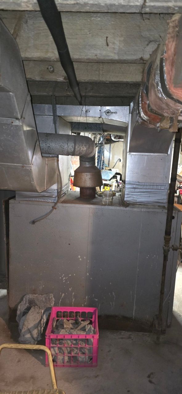 A grey metal furnace in a basement with silver ductwork, an exhaust pipe, and a pink crate on the floor in front of it.