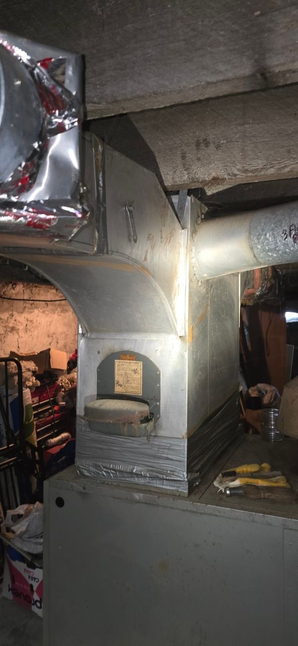 A close-up of a metal HVAC furnace plenum with attached ducts and a rectangular access panel in a basement setting.