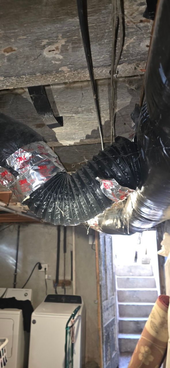 A black flexible HVAC duct curves and connects to a main duct pipe above a basement laundry area with stairs in the back.